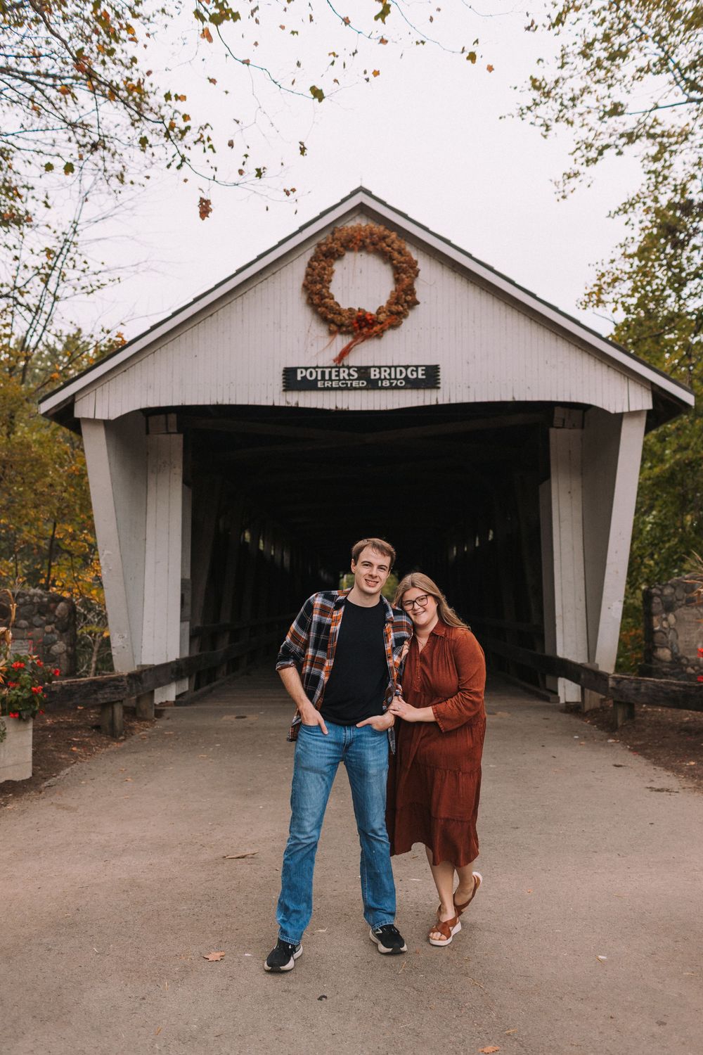 Couple poses playfully in front of a rustic covered bridge during autumn.