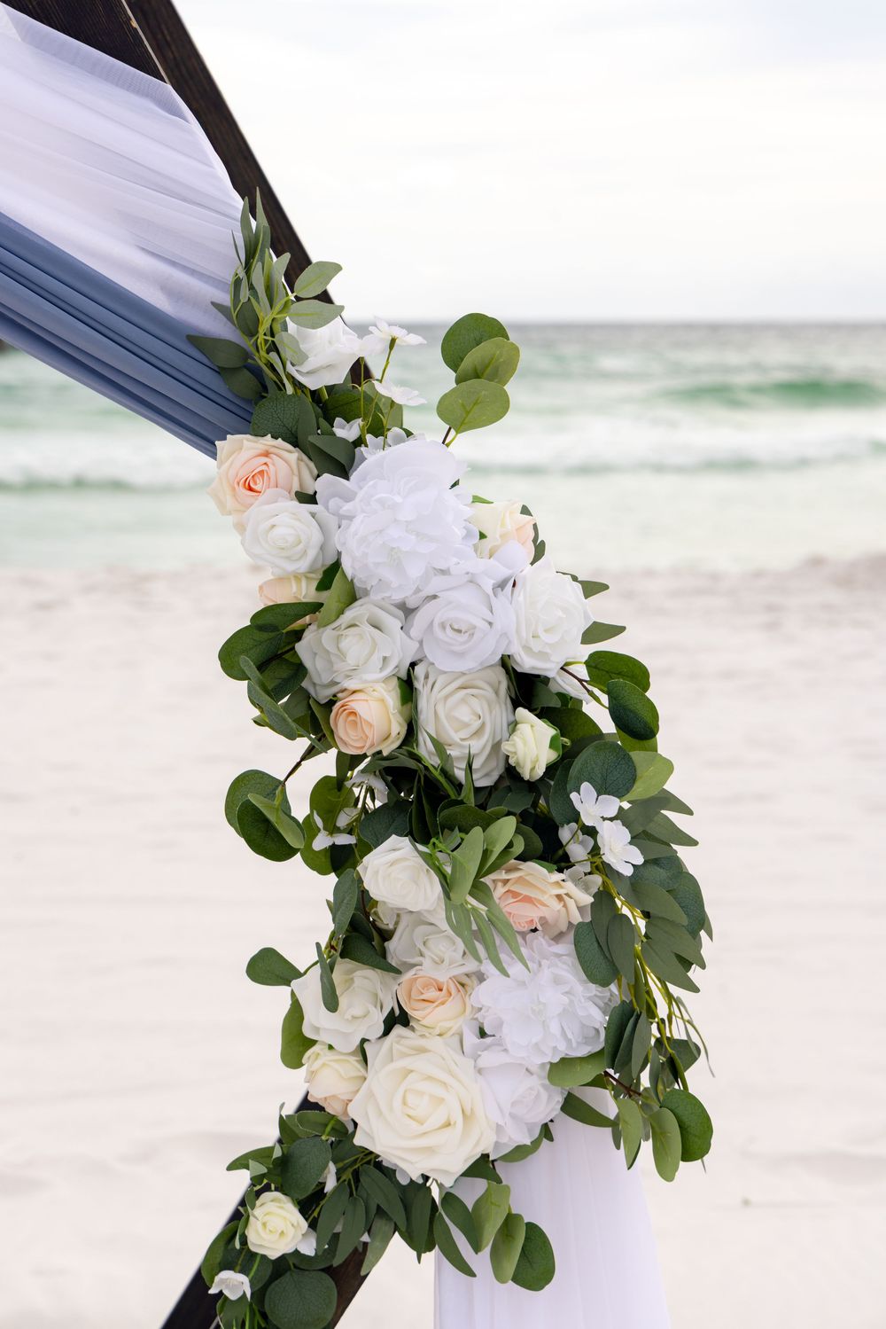 A romantic beach wedding arch decorated with white roses, peach flowers and eucalyptus greenery against ocean waves.