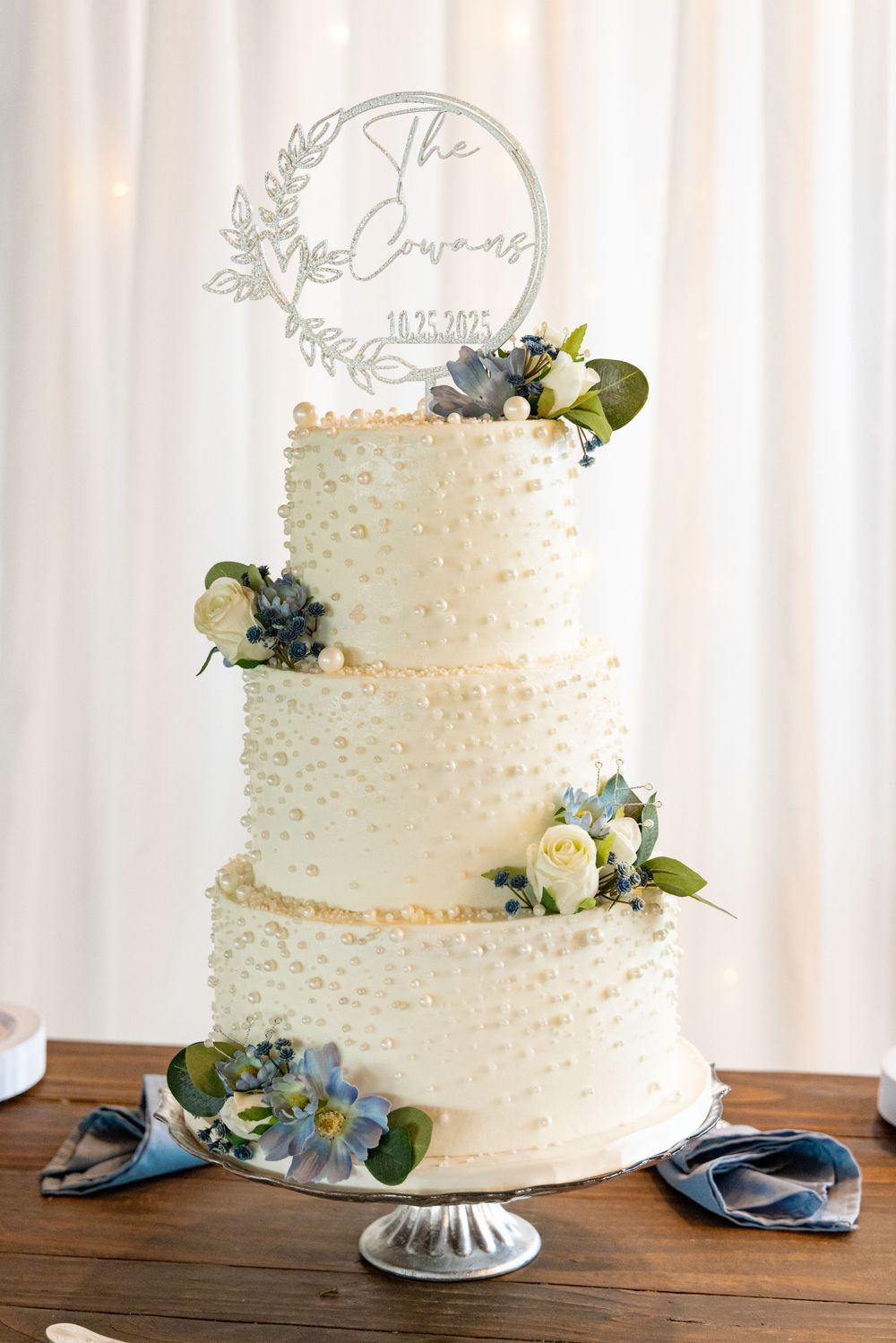 Three-tiered white wedding cake decorated with delicate dots and blue floral accents on a wooden table.