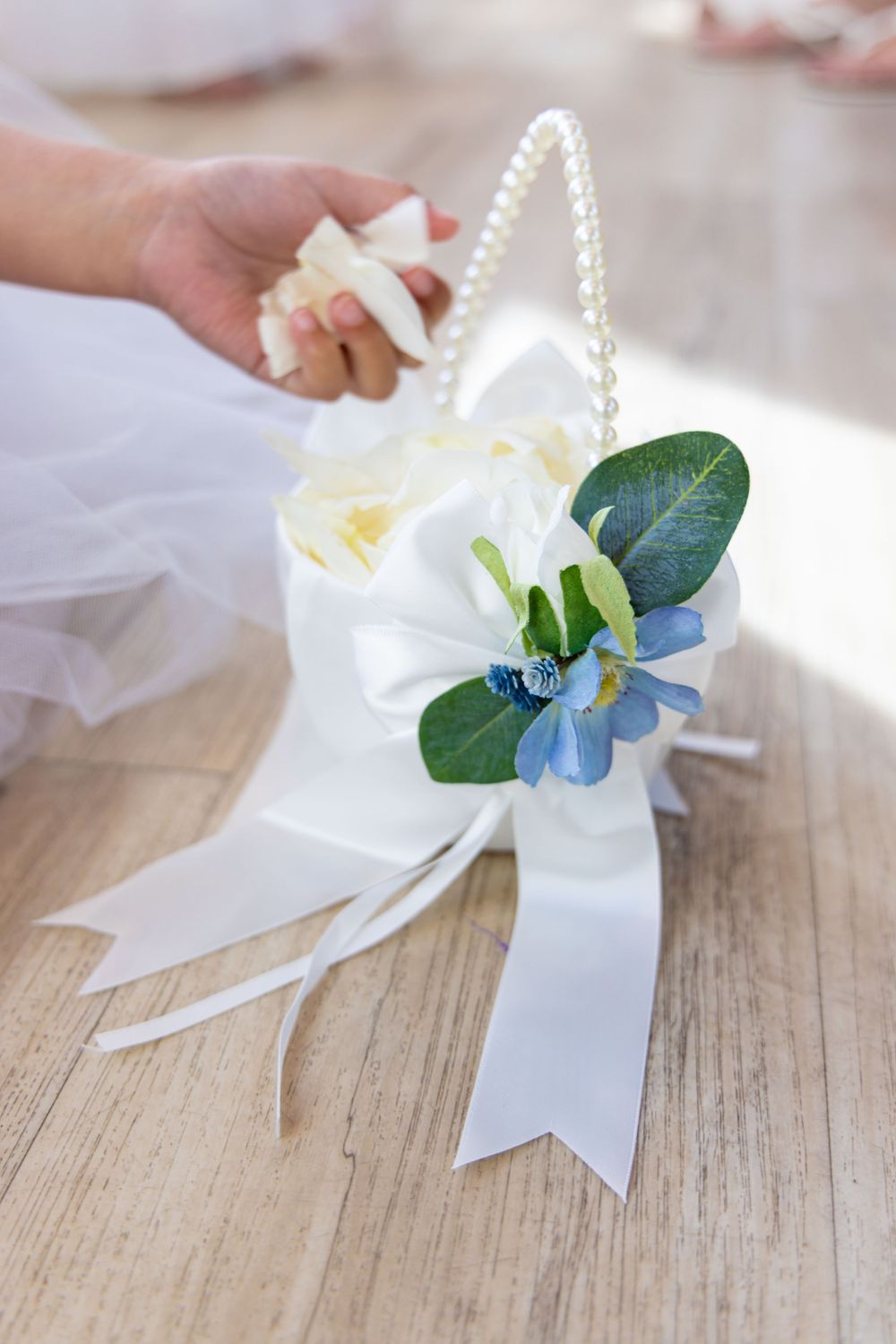 White satin bow with delicate blue flower and greenery being arranged on a rustic wooden surface.