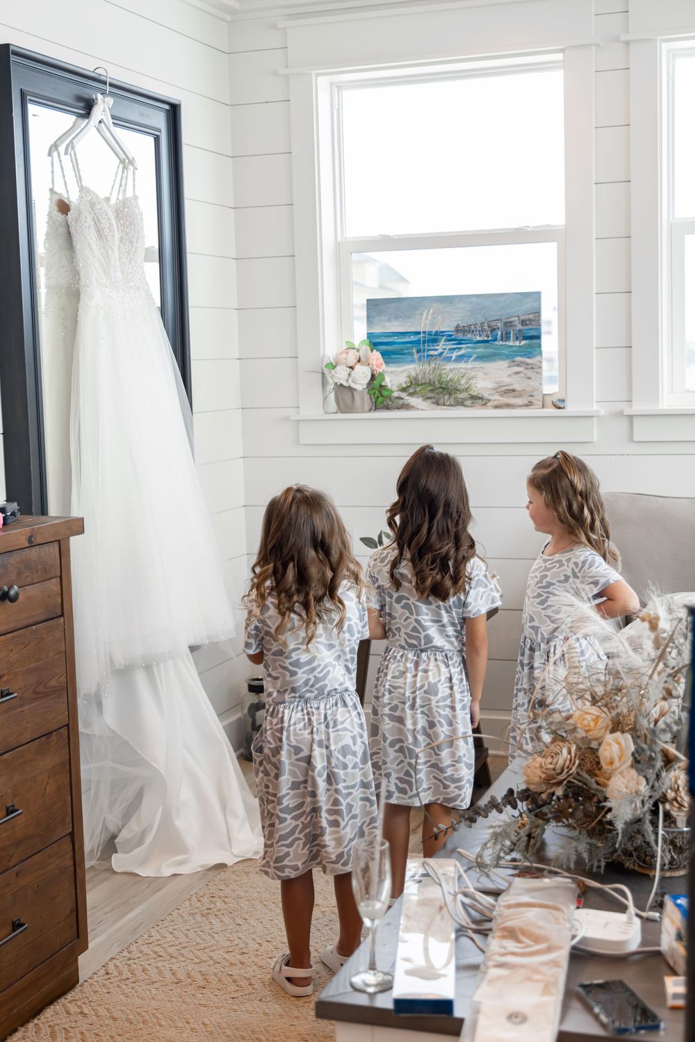 Three young attendants in floral dresses admire a white wedding gown hanging in a bright coastal-themed room.