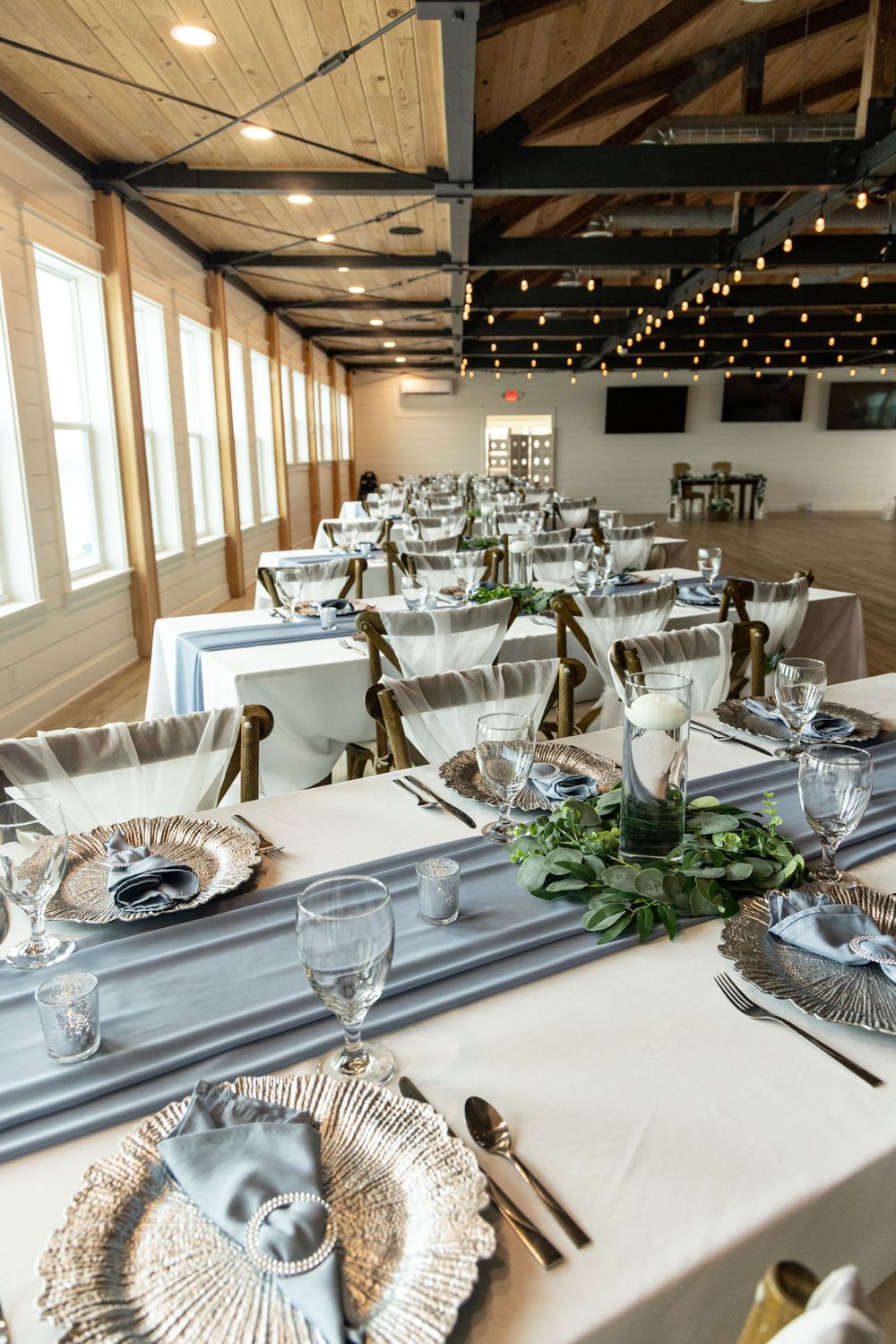 Elegant wedding reception tables set with white linens, gray runners, and greenery in a rustic venue with exposed beams.