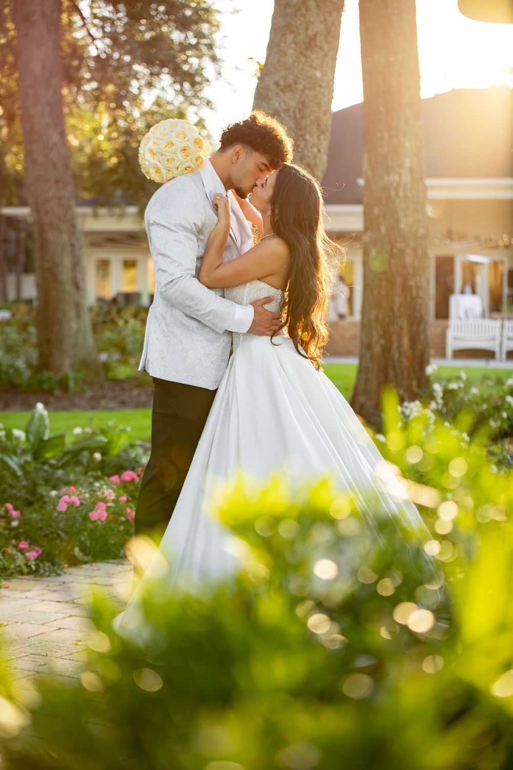 A romantic wedding couple shares a kiss at sunset in a garden setting with golden sunlight filtering through trees.