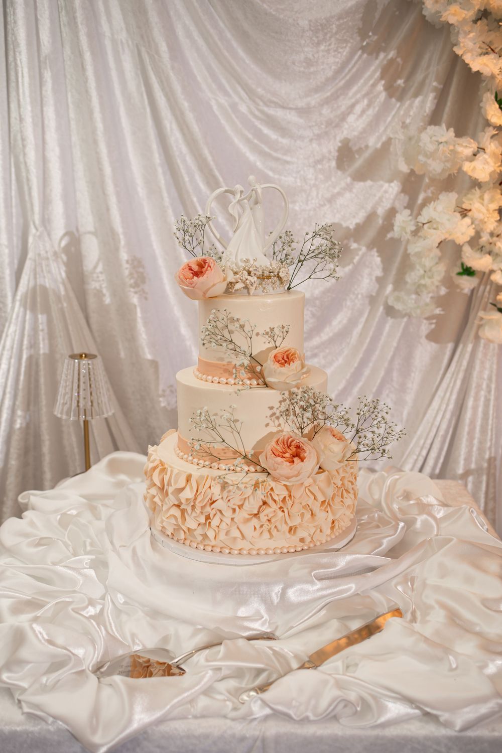 A delicate peach-colored wedding cake decorated with floral accents sits on white draped fabric with soft lighting.