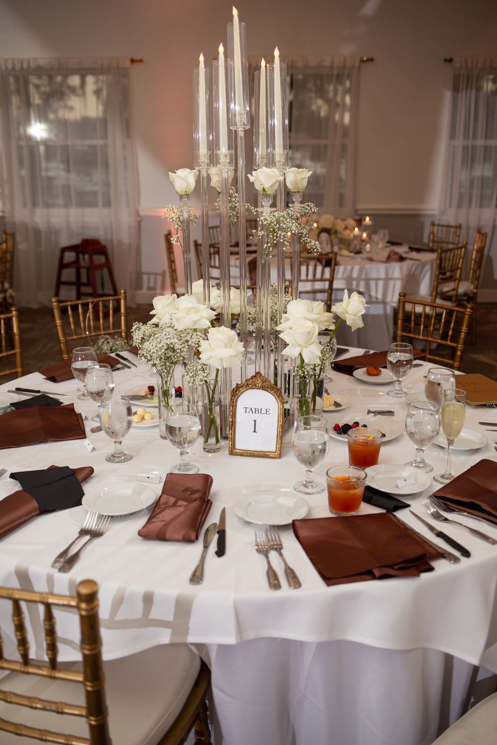 Elegant wedding reception tables with white linens, gold chairs, tall candelabras, and burgundy napkins set for formal dining.