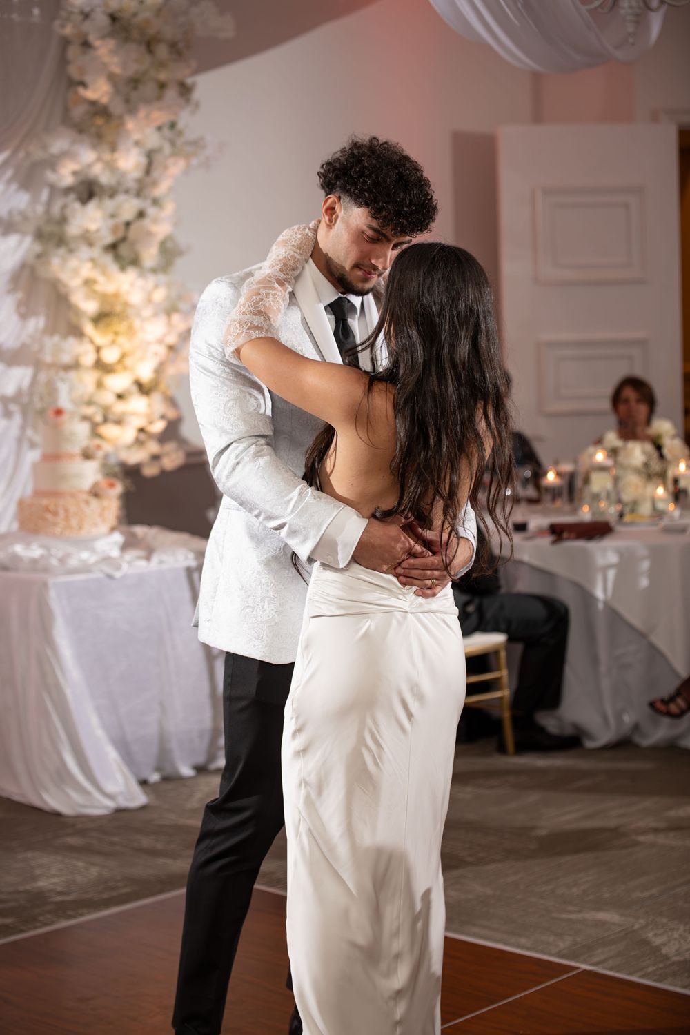 Romantic couple sharing an intimate dance moment on a wooden floor with white floral decorations in the background.