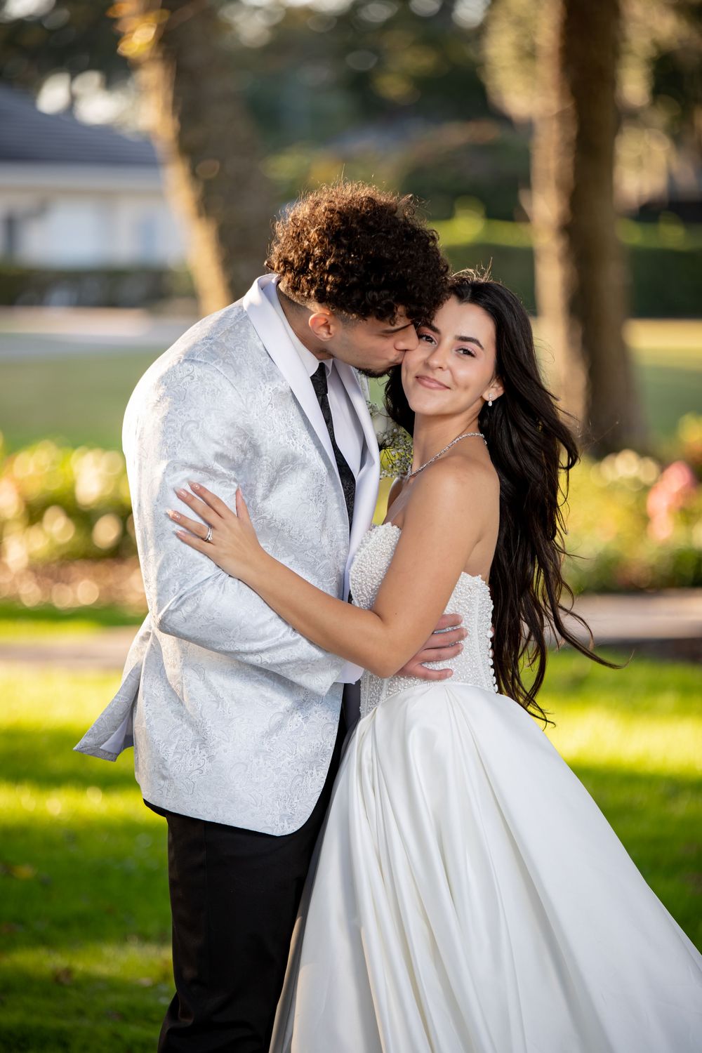 A romantic wedding couple embraces and dances in a sunlit garden setting with trees and flowers in the background.