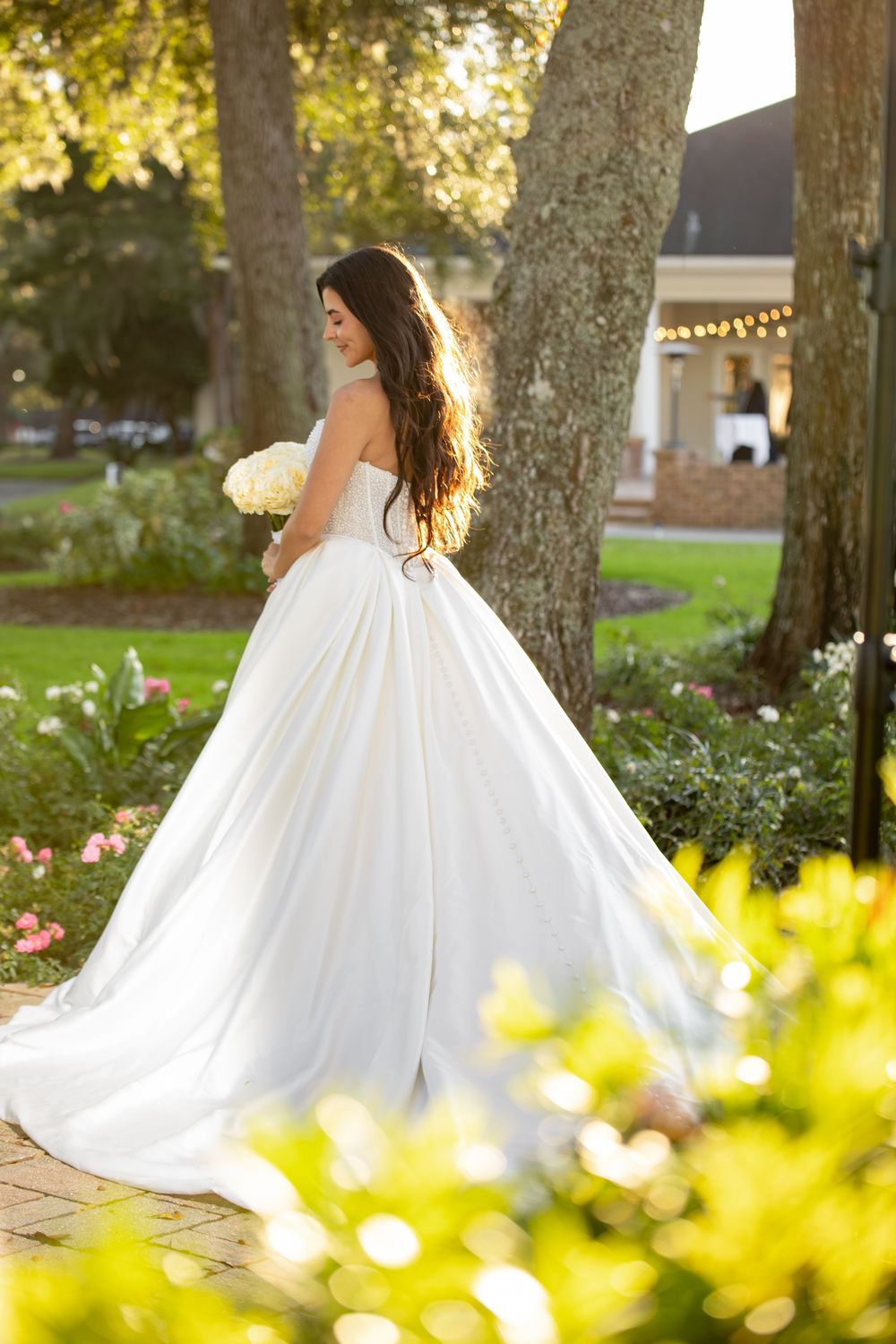 A flowing white wedding dress twirls gracefully in an outdoor garden setting with sunlit trees in the background.