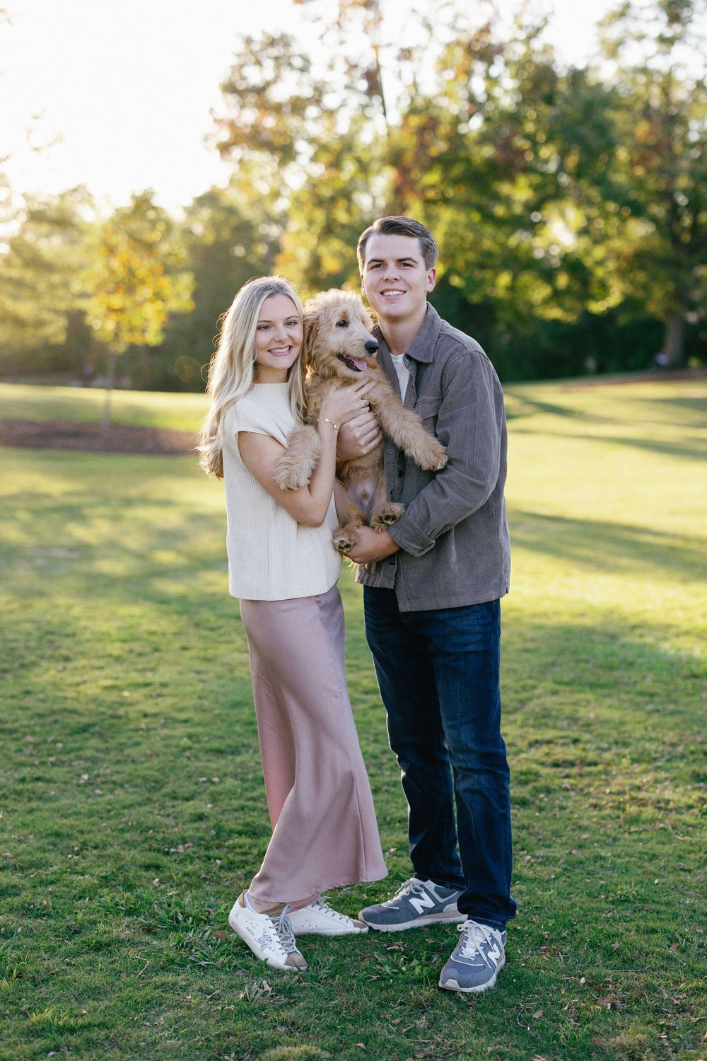 A couple poses with their golden retriever puppy in a sunny park during golden hour.