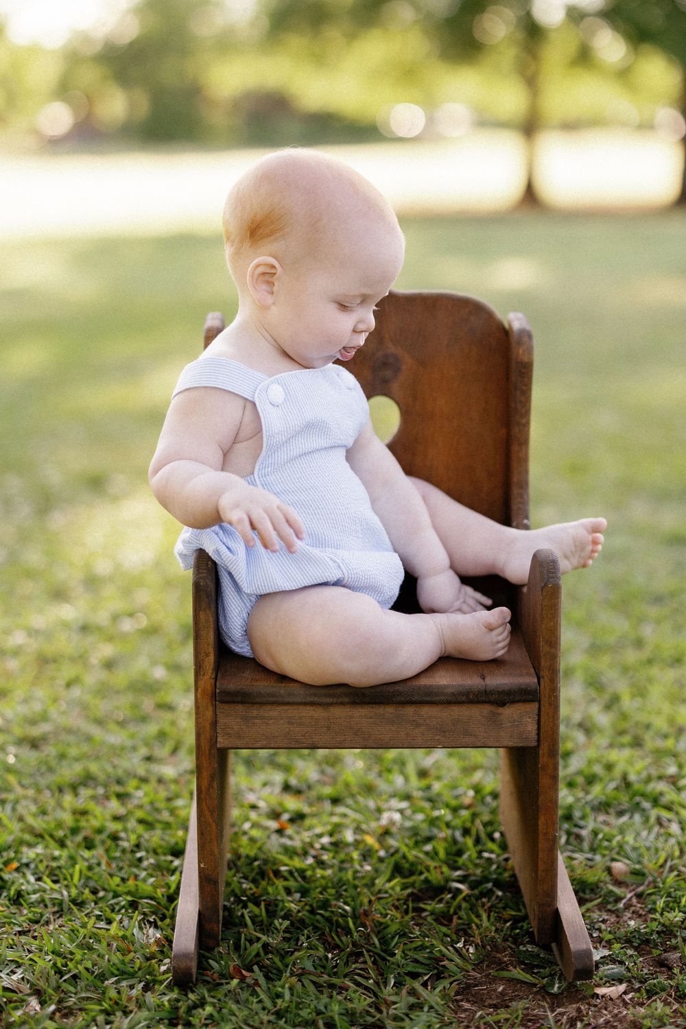 Baby in white romper sits on small wooden rocking chair outdoors on grass with sunlit trees in background.