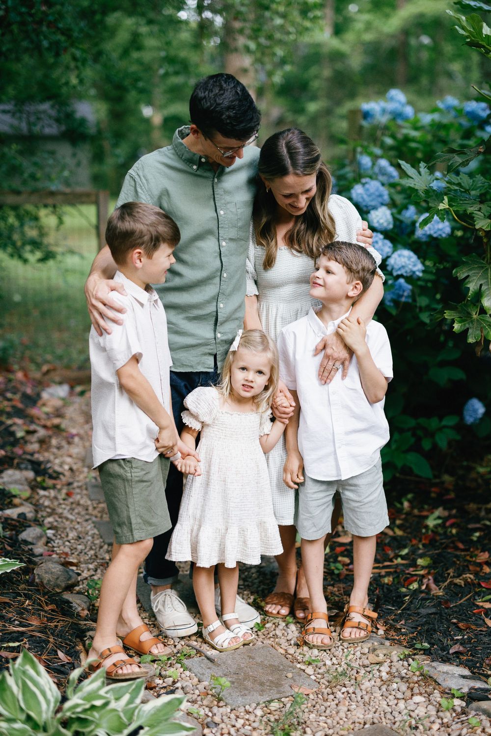 A family in coordinated white and grey outfits poses together in a garden setting with blue hydrangeas and stone path.