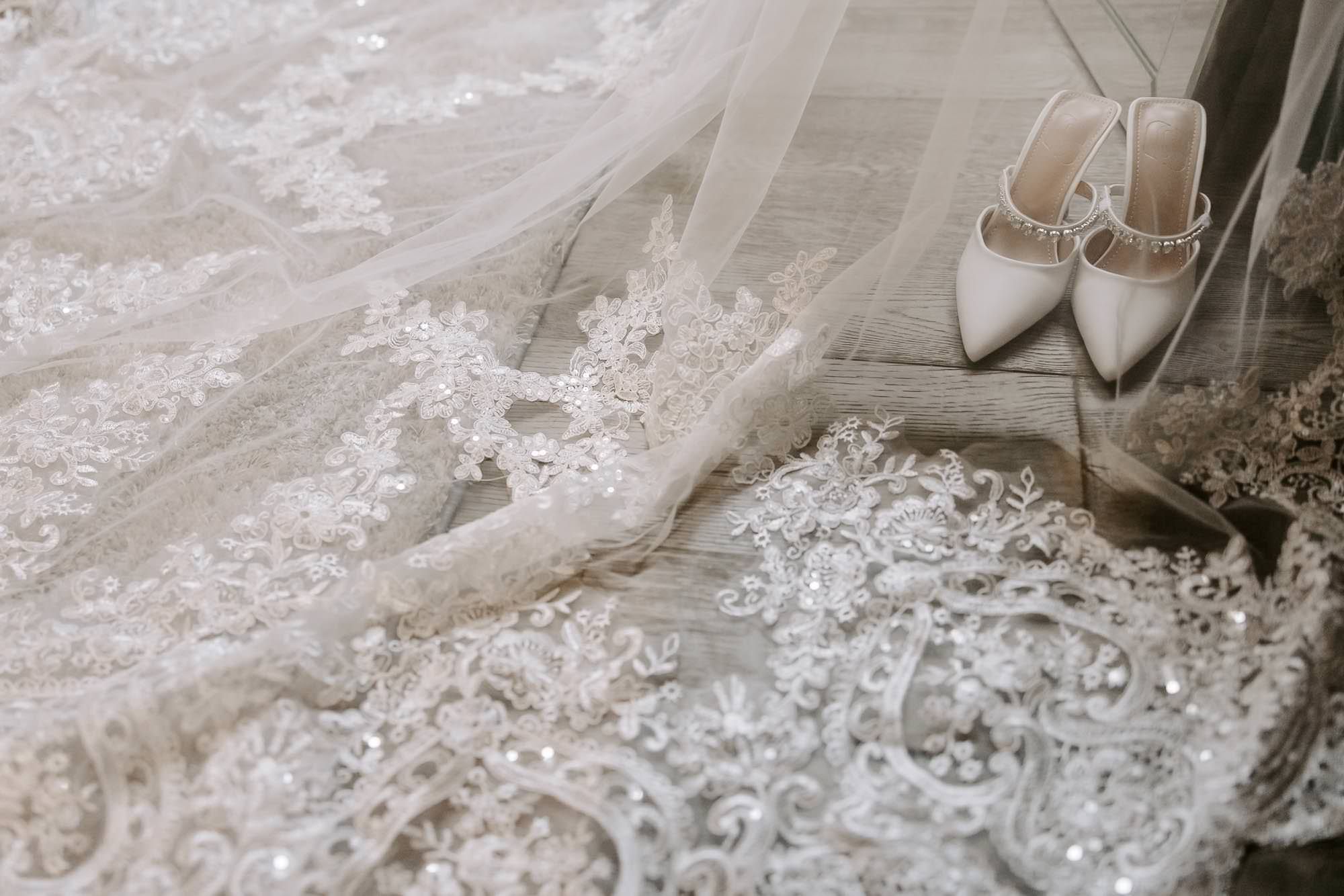 A delicate white lace wedding dress hangs from a hanger, showcasing intricate floral embroidery and beaded details.