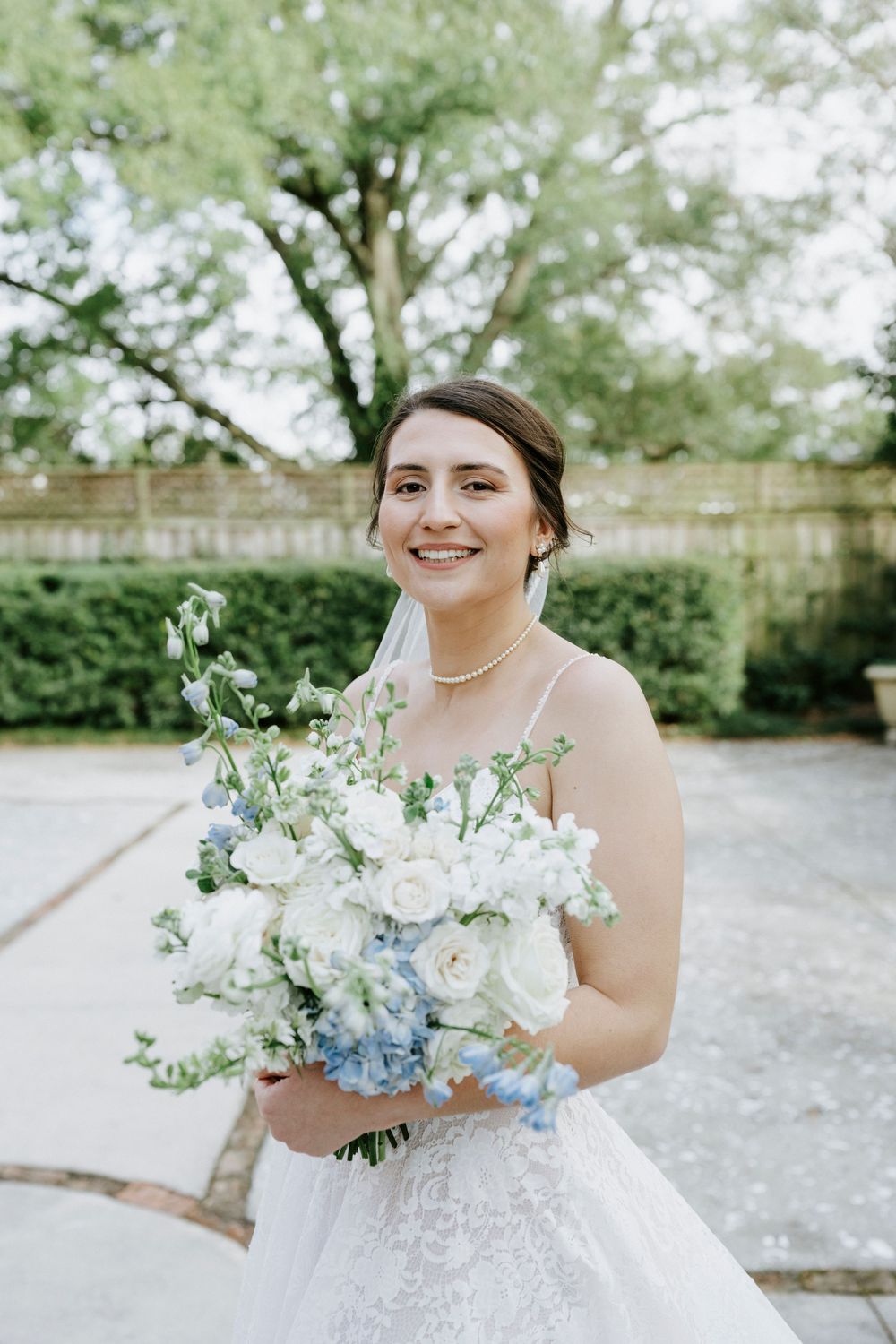 A bride in a white lace wedding dress holds a beautiful bouquet of white and pale blue flowers in an outdoor garden setting.