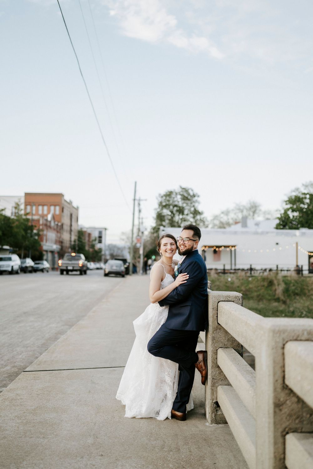 Couple in wedding attire sits together on a concrete ledge overlooking an urban landscape on their special day.