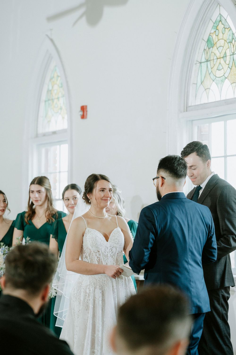 A wedding ceremony takes place in a bright chapel with stained glass windows and emerald green bridesmaid dresses.
