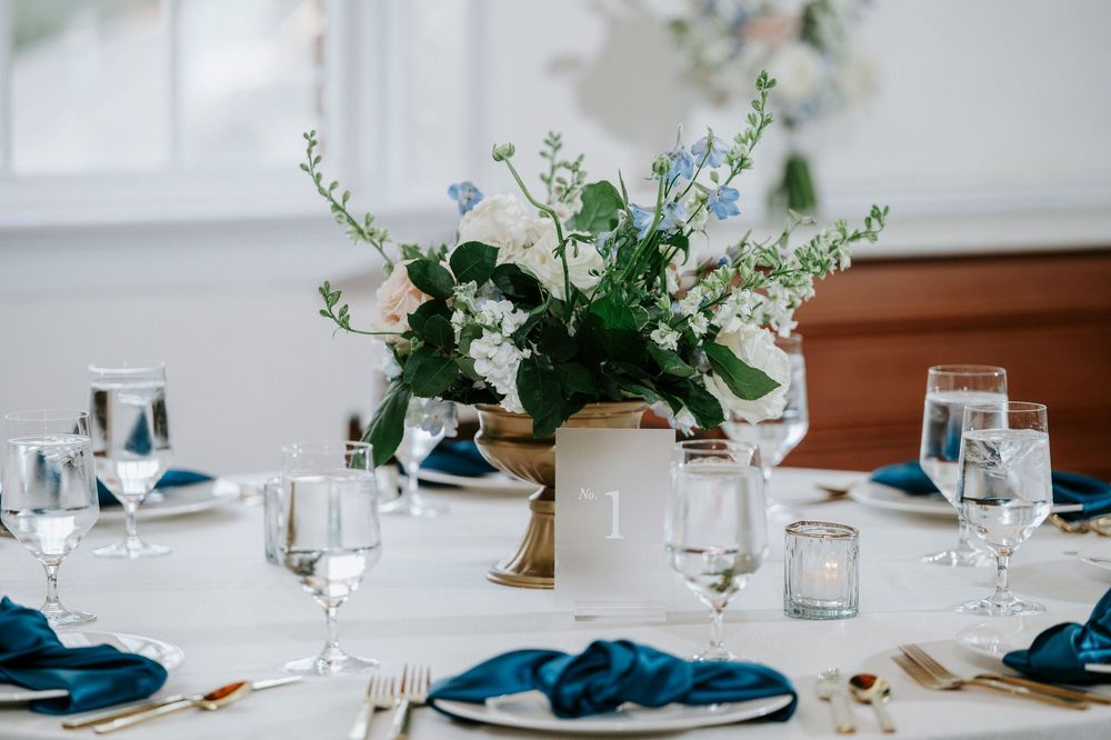 Elegant table setting with white ceramic vase centerpiece filled with greenery and navy blue napkins on white tablecloth.