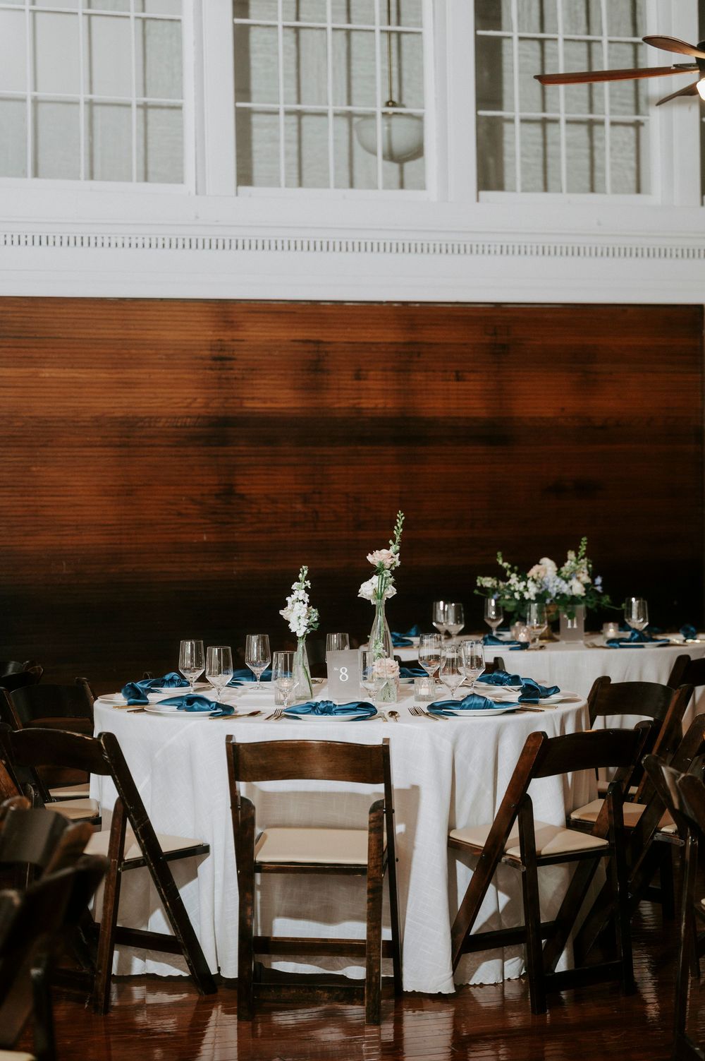 Elegant wedding reception venue with round tables set with white linens, navy blue accents, and rustic wooden chairs.