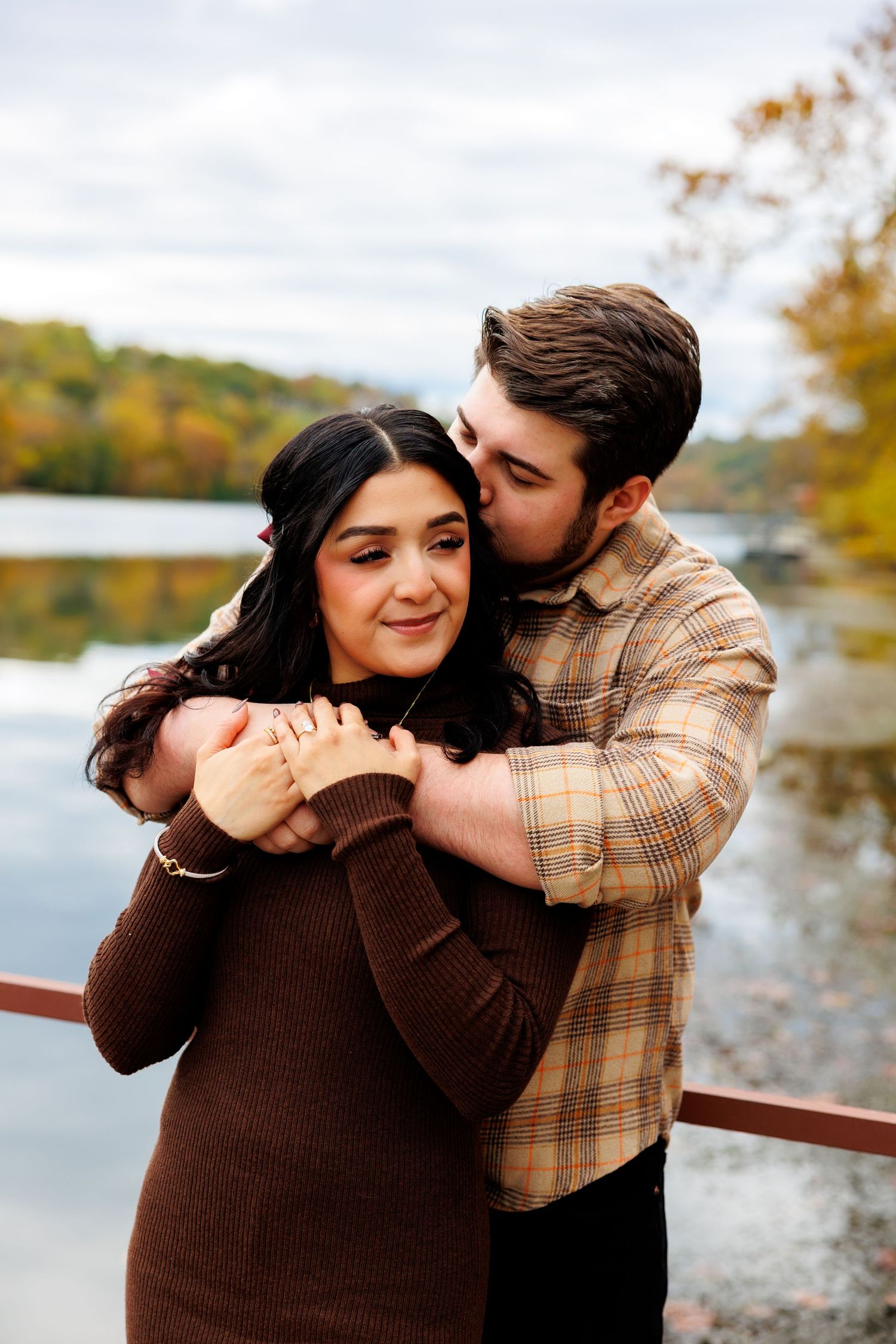 A couple stands together by a serene lake surrounded by autumn trees and mountains in the background.