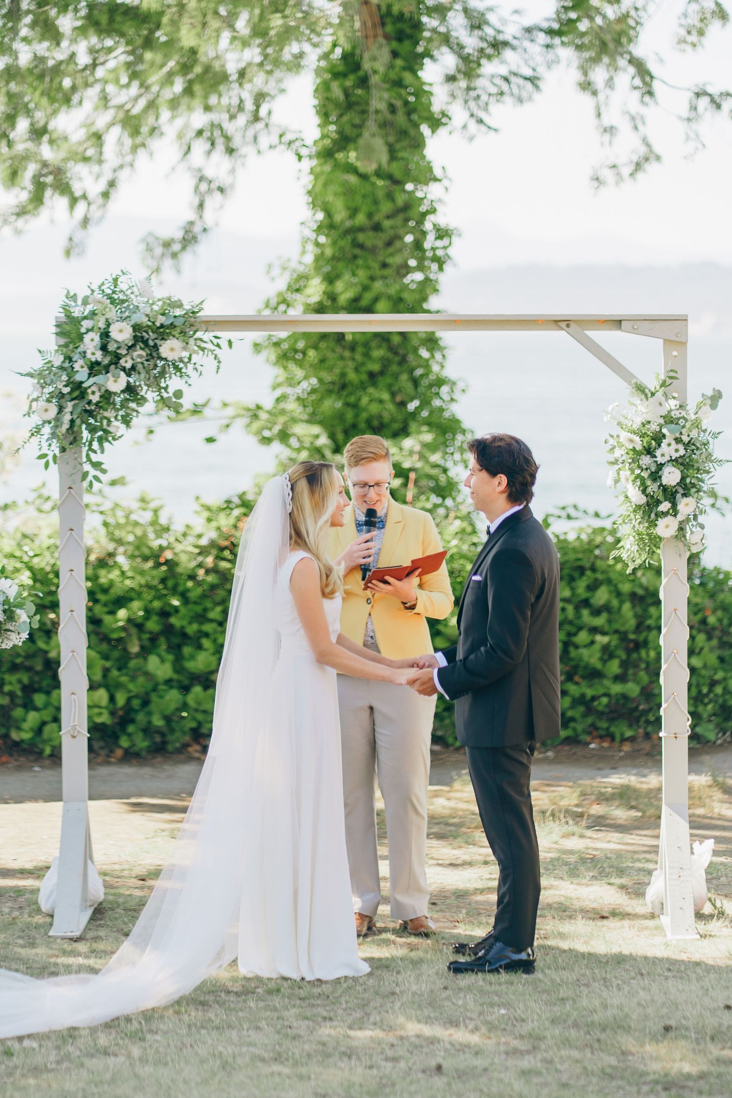 An outdoor wedding ceremony takes place under a white pergola with greenery and trees in the background.