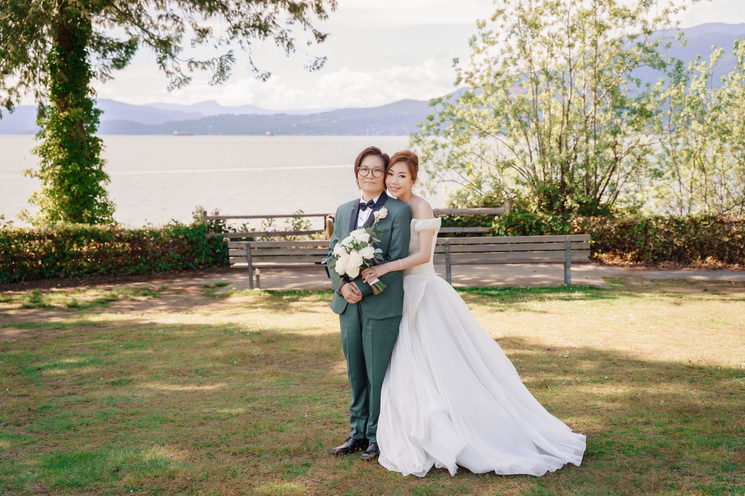 A couple in wedding attire poses together on a grassy lawn overlooking a scenic lake and mountains.