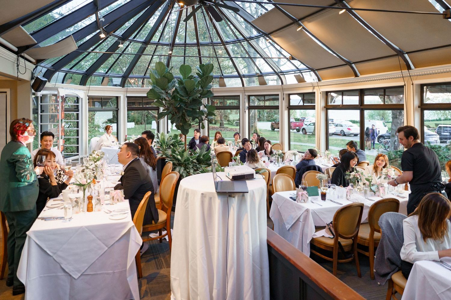 Elegant restaurant dining room with glass atrium ceiling and white tablecloths filled with patrons enjoying their meals.