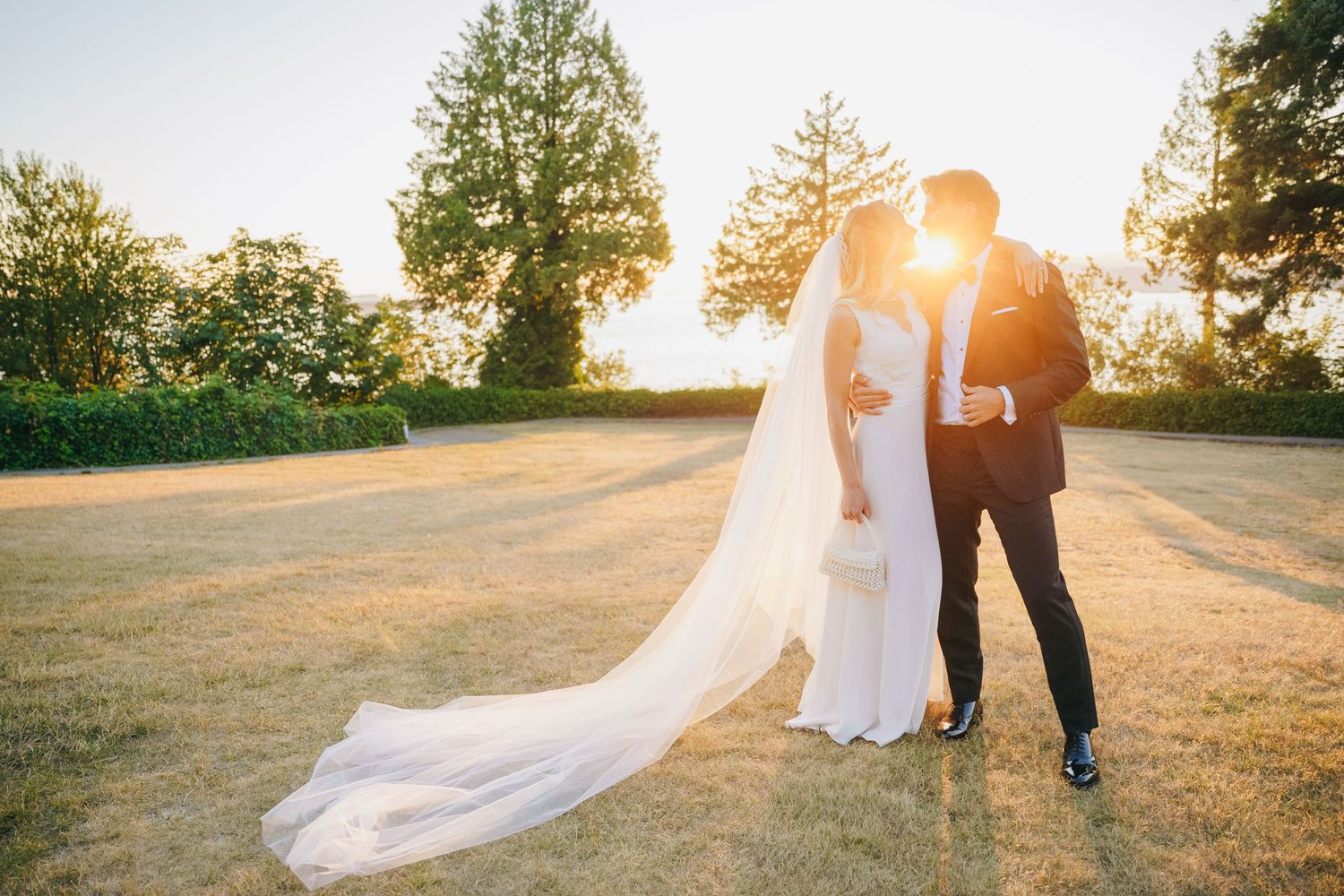 A couple in wedding attire stands together in a sunlit field during golden hour as a veil flows in the breeze.