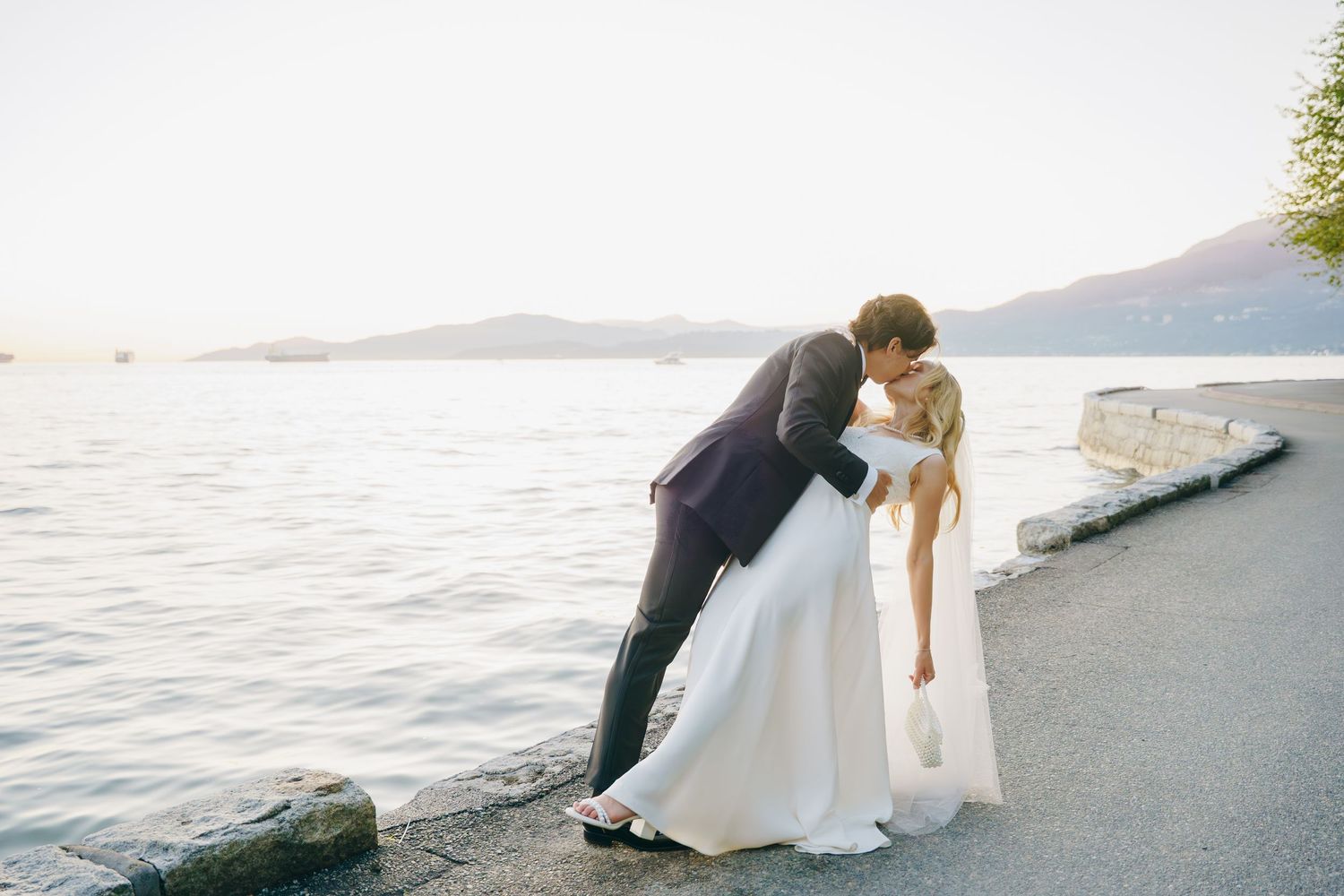 A romantic couple in wedding attire poses along the waterfront at sunset with mountains in the distance.