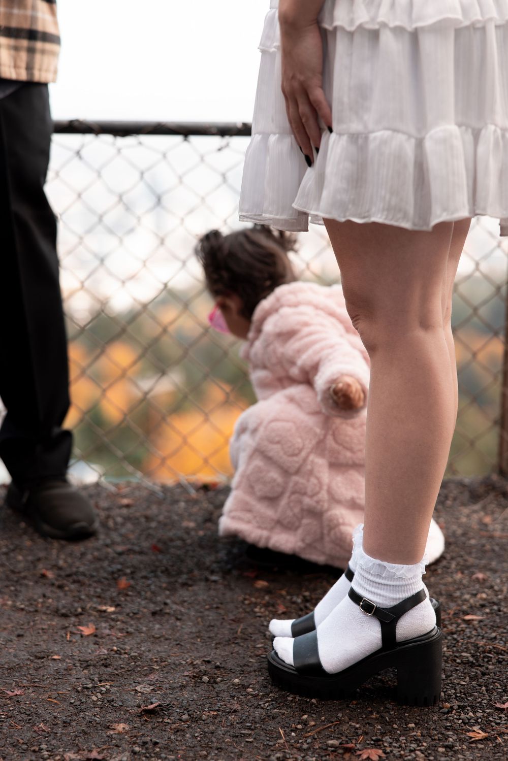 A child's feet wearing white socks and black mary jane shoes stand on dark soil next to a pink plush toy.