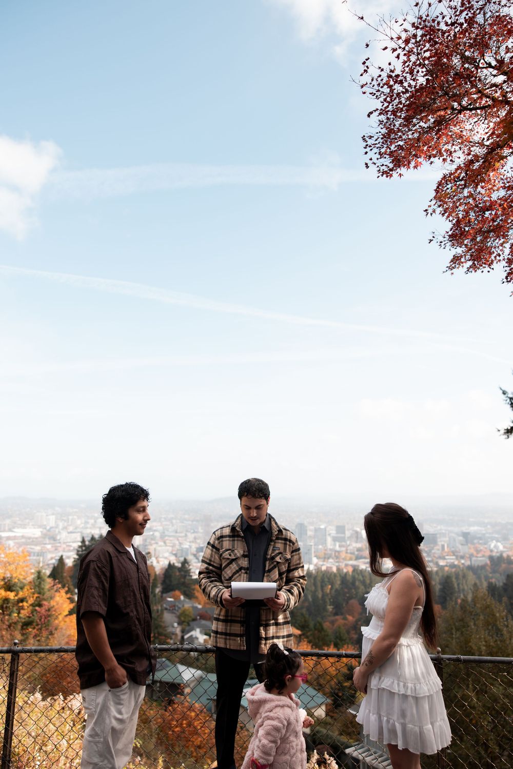 A couple shares a romantic moment at a scenic overlook with autumn trees and city views in the background.