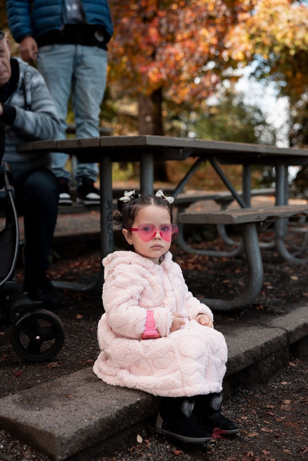 A baby wearing a pink dress and sunglasses sits on outdoor steps during autumn with colorful fall foliage in background.