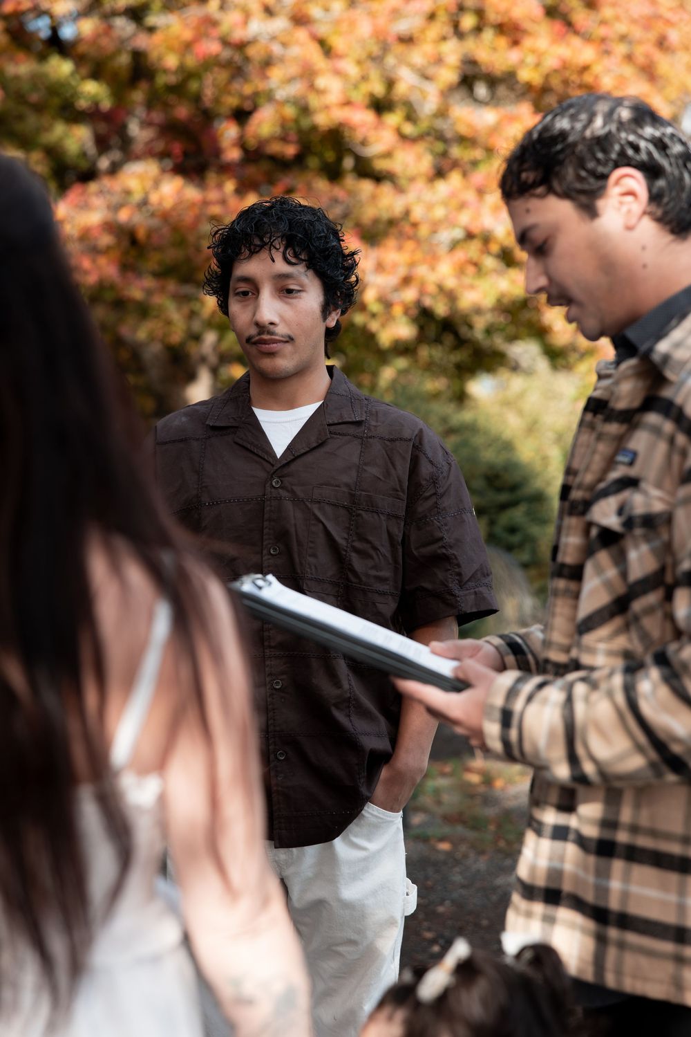 Two students chat while holding books on a path surrounded by autumn foliage with orange and yellow leaves.