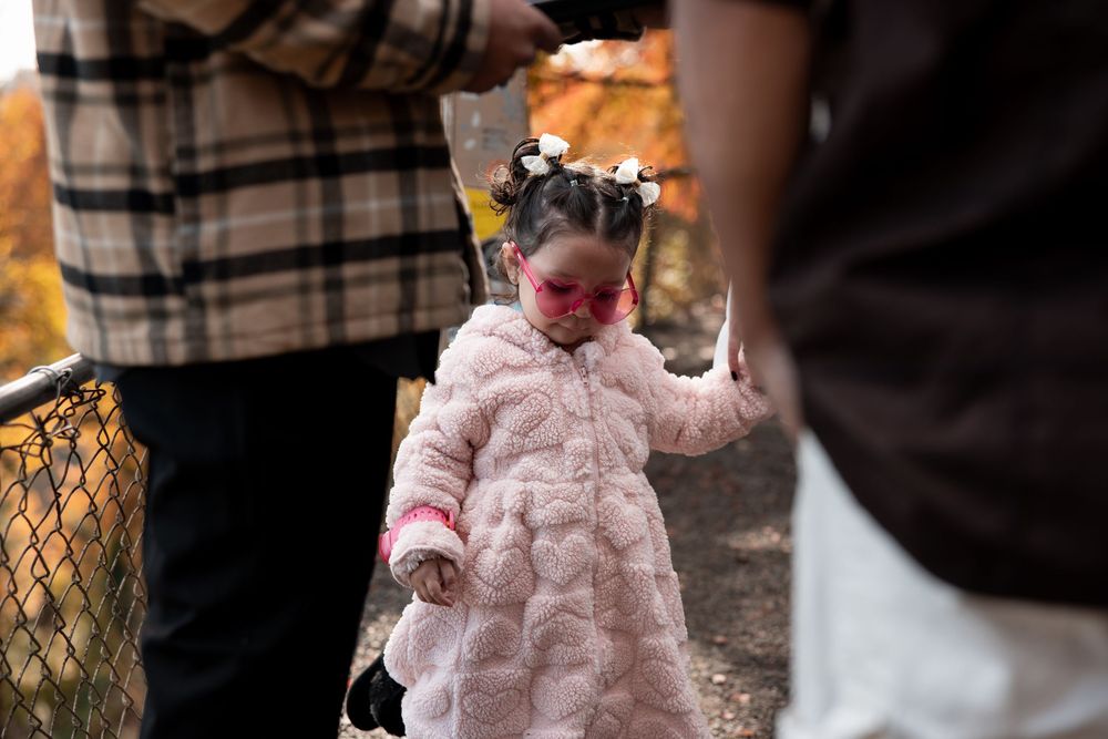 A child wearing a pink winter coat and tiara walks between adults during an outdoor autumn gathering.