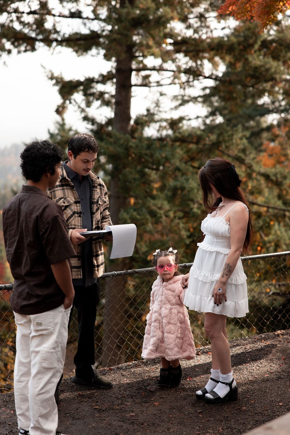 A group gathers outdoors in formal attire during autumn, with evergreen trees and fall foliage in the background.