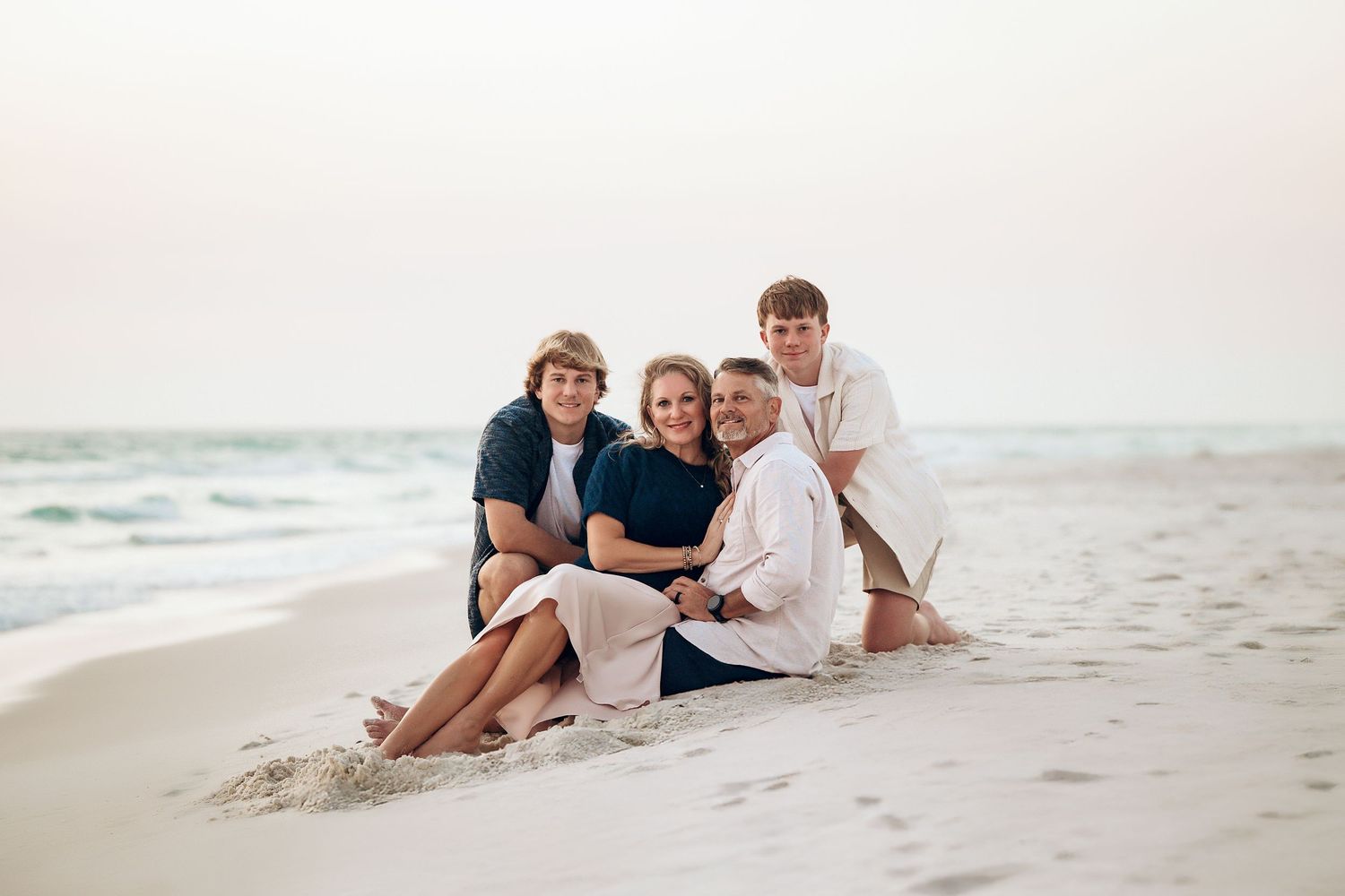 A family wearing white and blue clothing sits together on a sandy beach during sunset.