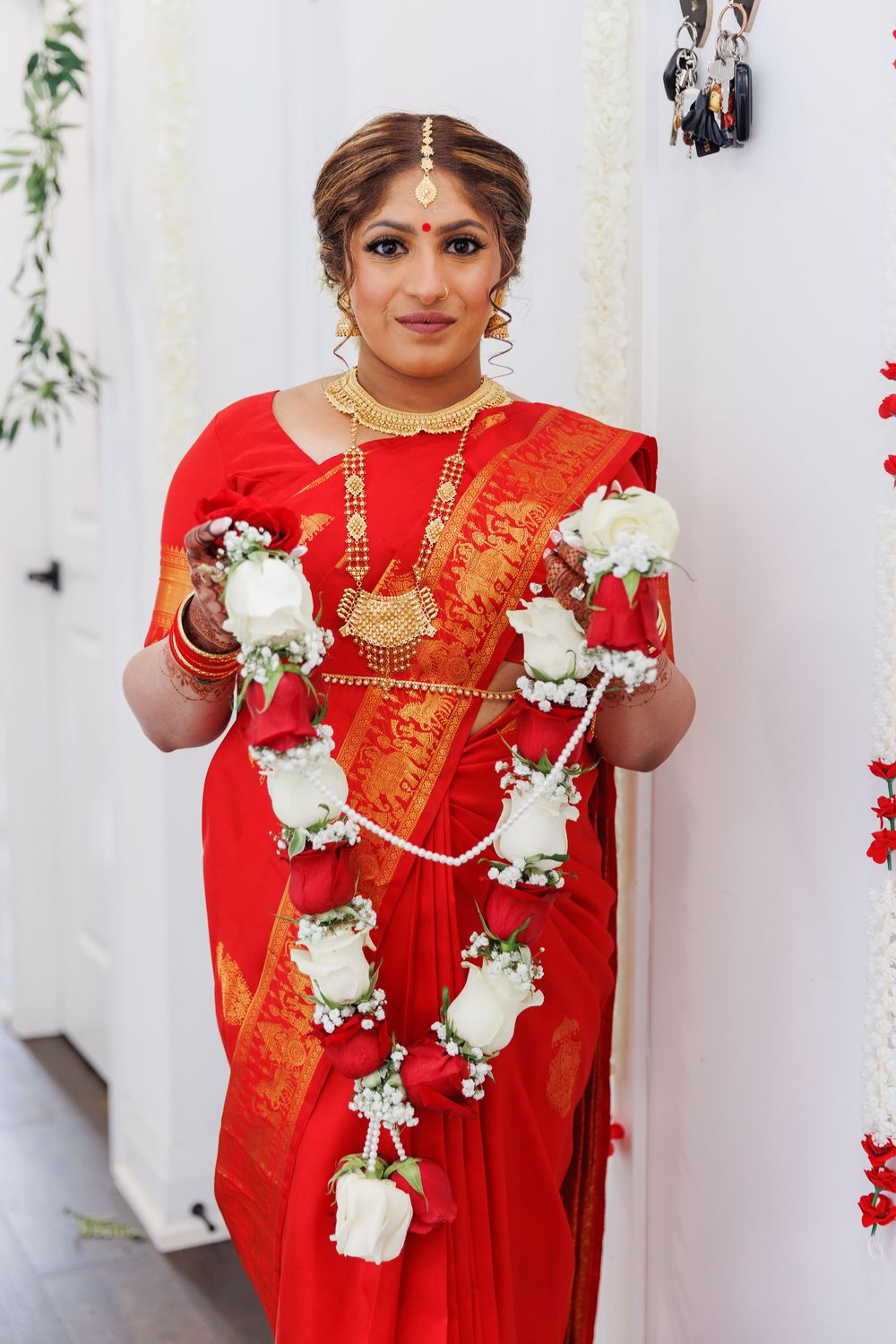 Traditional South Asian bride wearing red silk saree and gold jewelry holding white floral garlands.