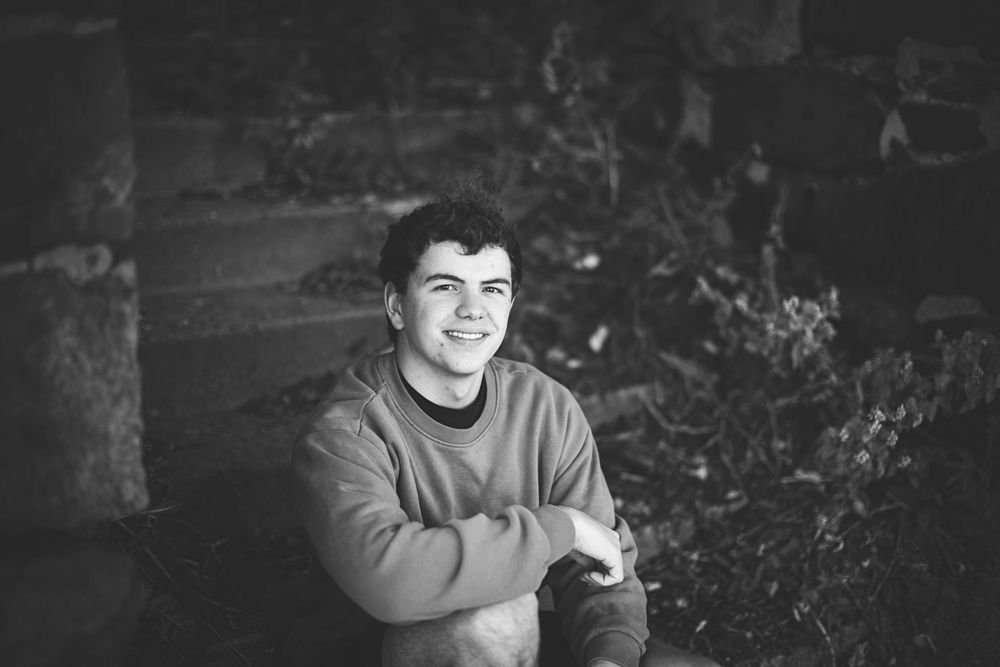 A black and white photograph shows a person in a sweater sitting outdoors and smiling at the camera.