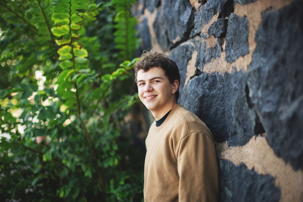 A person in a tan sweater smiles while standing against a stone wall with green foliage visible in the background.