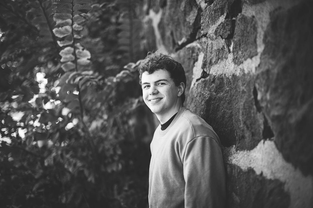A person smiles while sitting outdoors against a tree trunk in a natural setting.