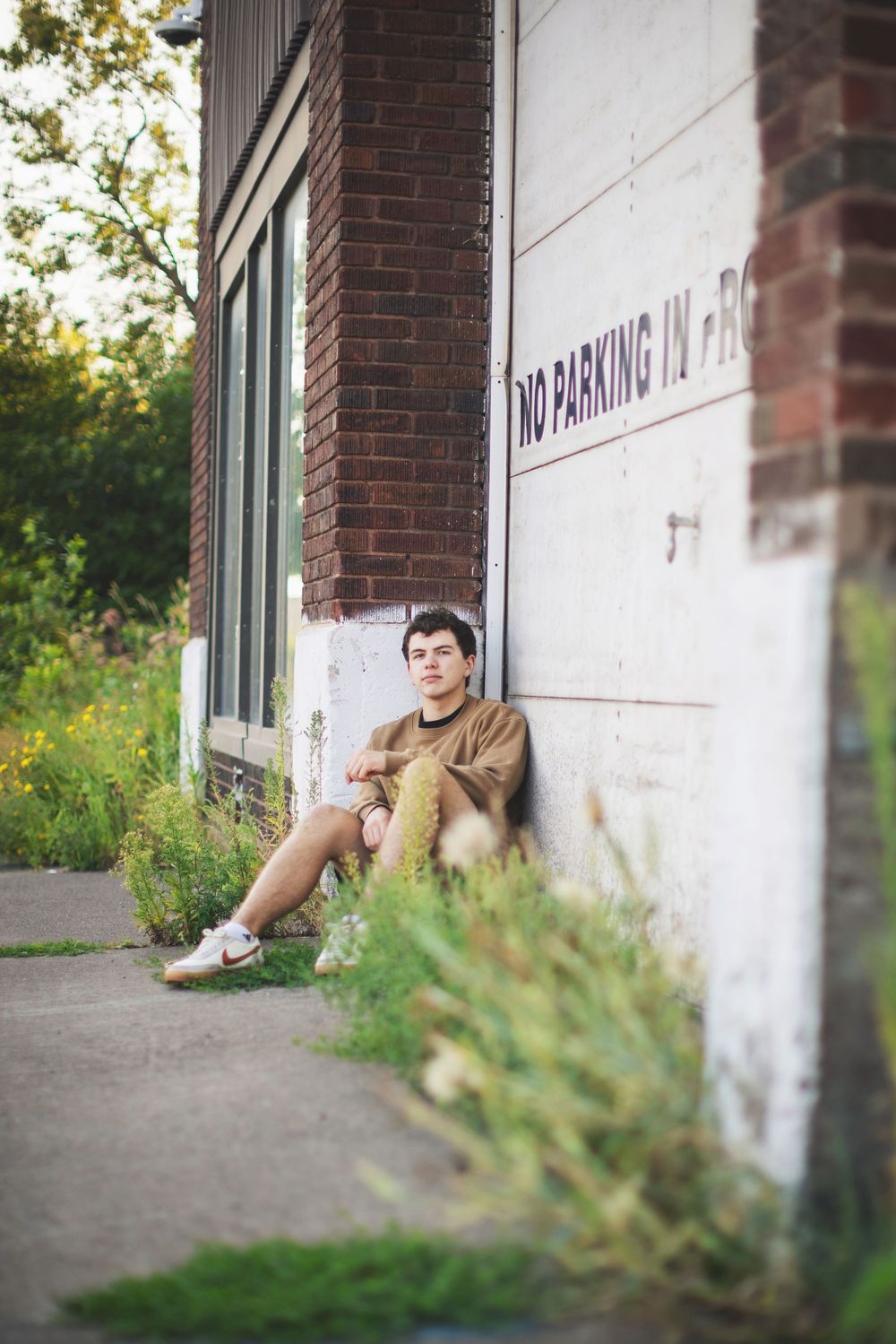 Someone sits casually against a brick wall with a 'No Parking' sign, wearing casual attire in an urban setting.