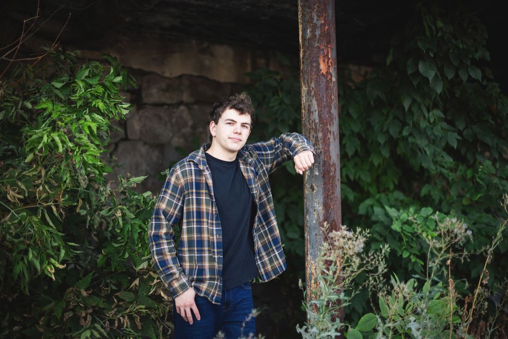Someone in a plaid shirt and jeans poses against a rustic wooden post surrounded by green foliage.
