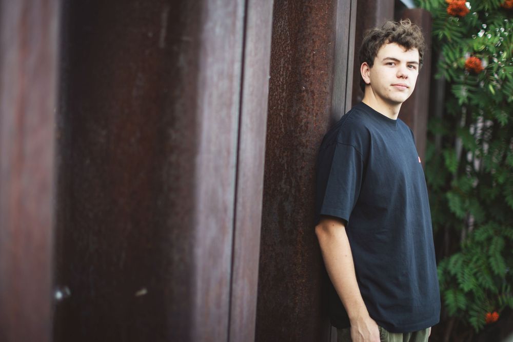 A young person in a black t-shirt stands against a dark brown wall with red flowers visible in the background.