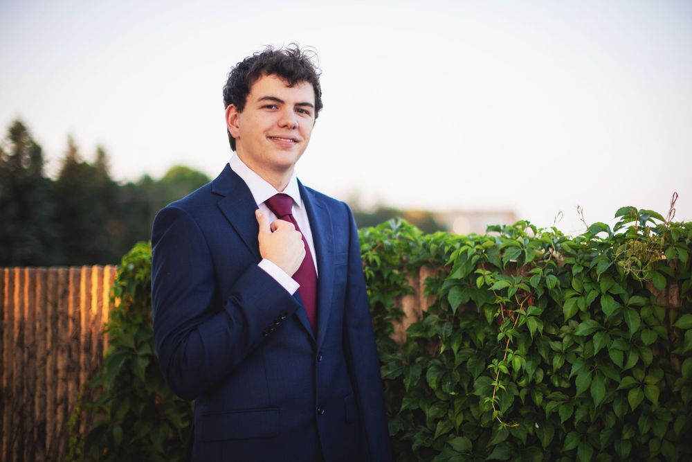 A person in a navy suit and red tie stands smiling outdoors against a green hedge backdrop.