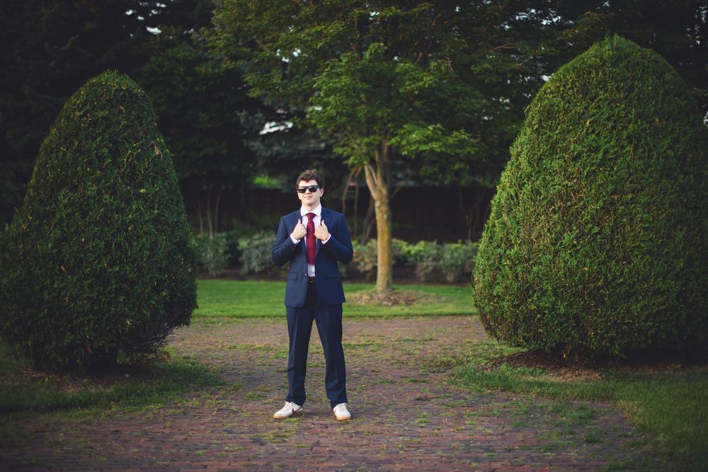 A person in a navy suit stands in a lush garden setting with manicured hedges and trees.