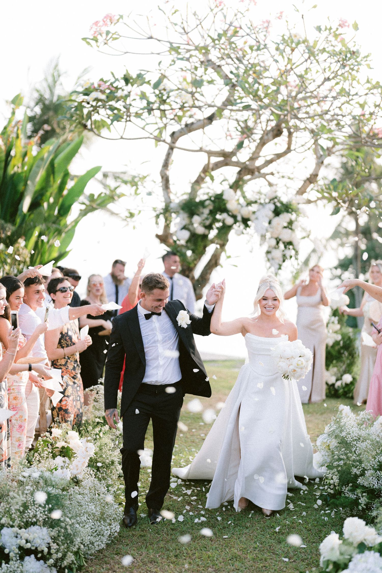 Wedding party celebrates outdoors with white flower petals being tossed in the air during a joyful garden ceremony.