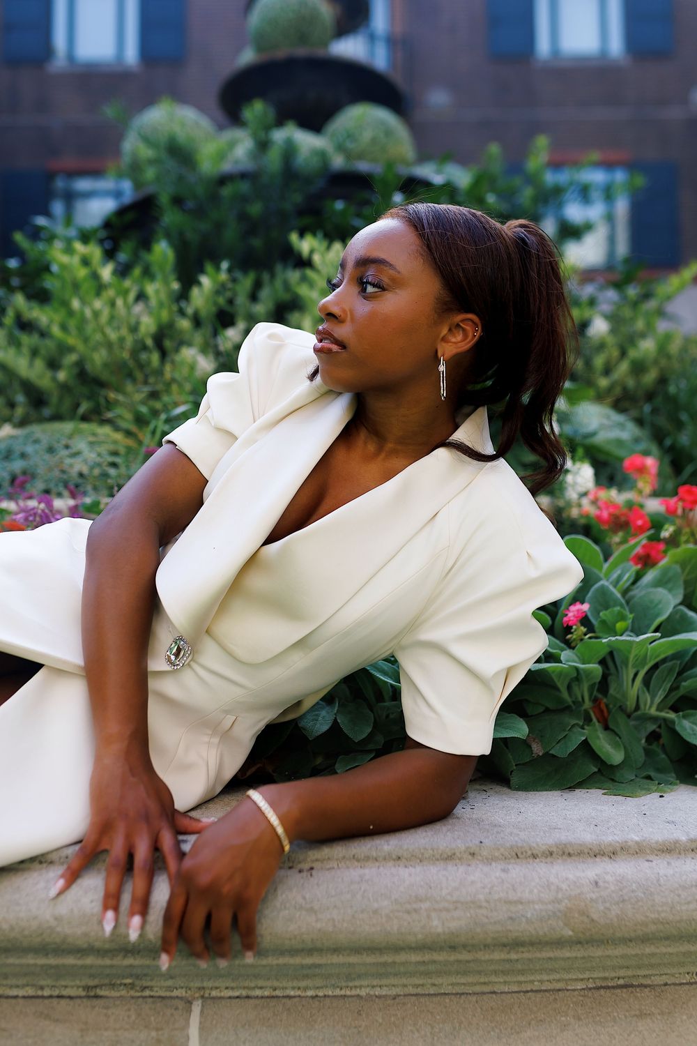 A person in a cream colored outfit lies on a concrete ledge in a garden with red flowers and greenery.