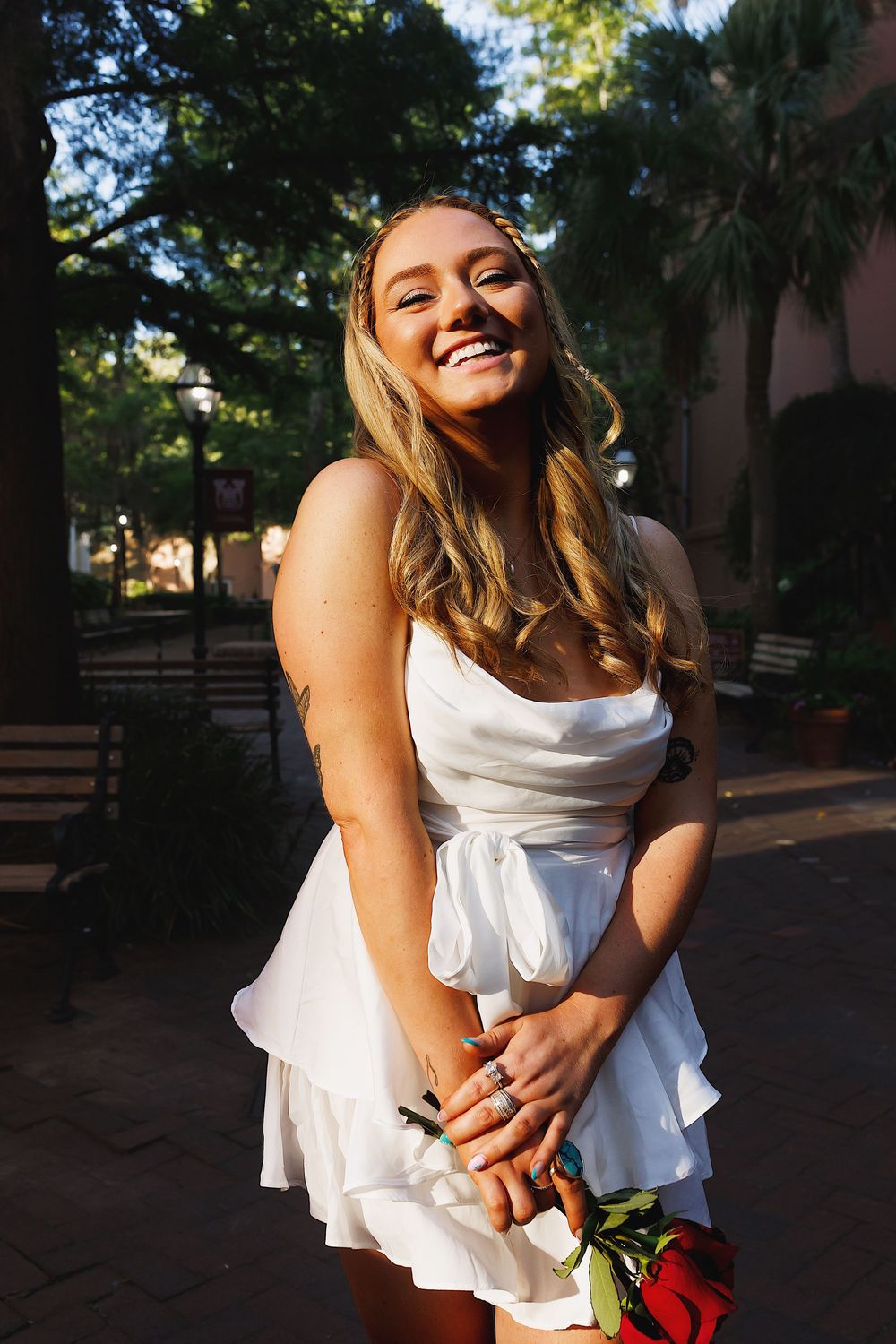 A person in a flowing white dress laughs joyfully while holding red roses in an outdoor garden setting.