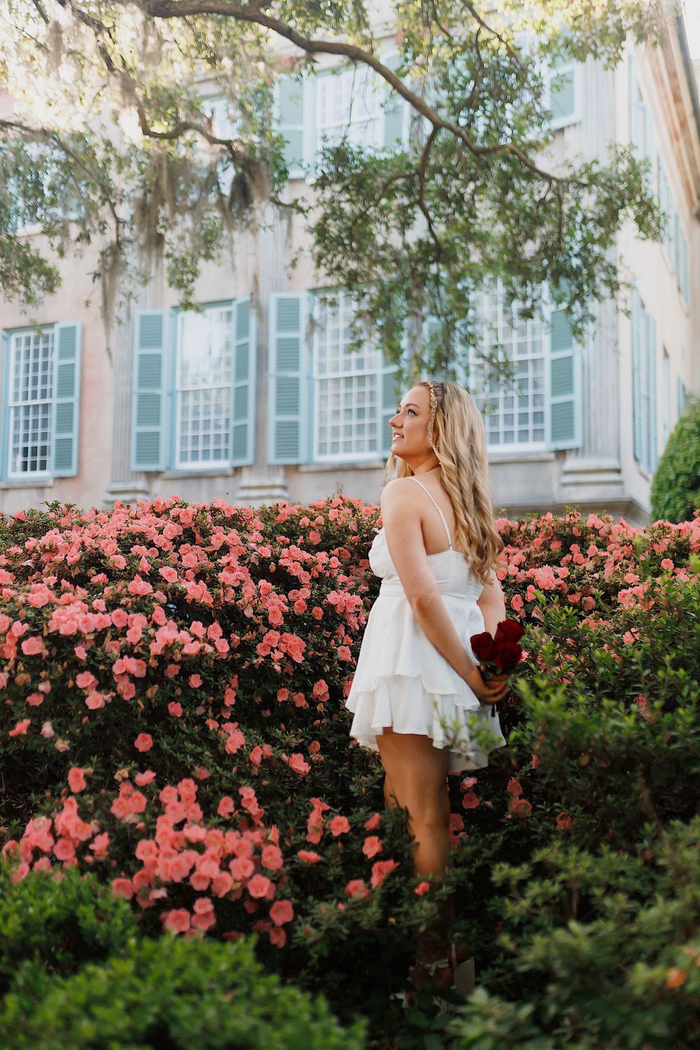 Woman in white dress stands among blooming pink azalea bushes beside a historic building with blue shutters.