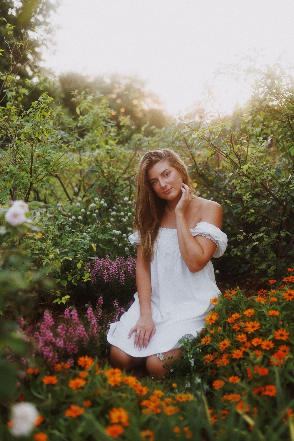 A woman in a white dress poses gracefully among vibrant orange marigolds and purple wildflowers at sunset.