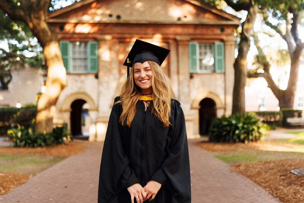 Graduate in cap and gown poses on tree-lined campus pathway during graduation photo session.