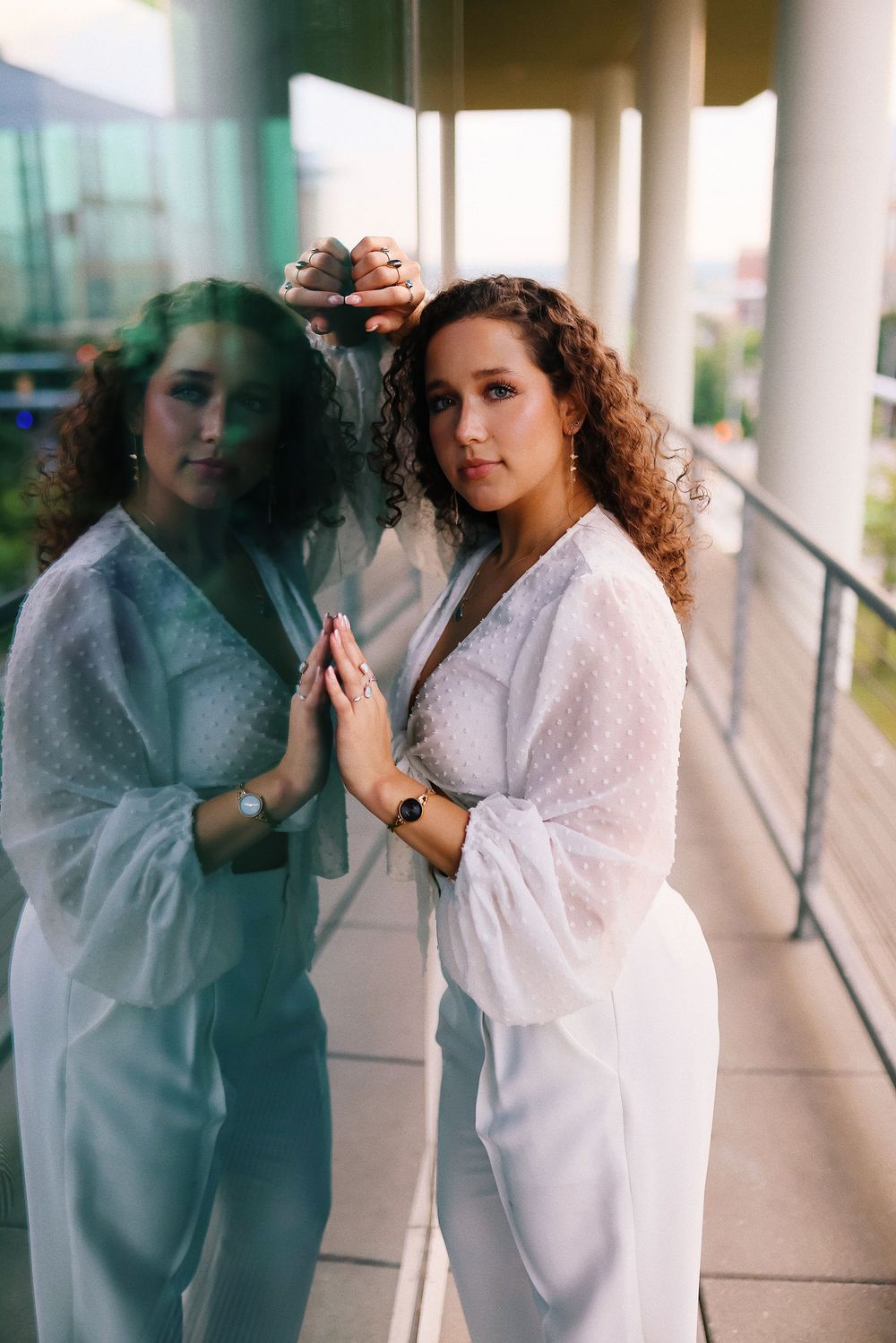 A person in white clothing poses next to their reflection in a modern glass window.